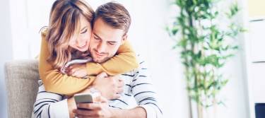 Frau umarmt Mann und schaut mit ihm gemeinsam aufs Smartphone